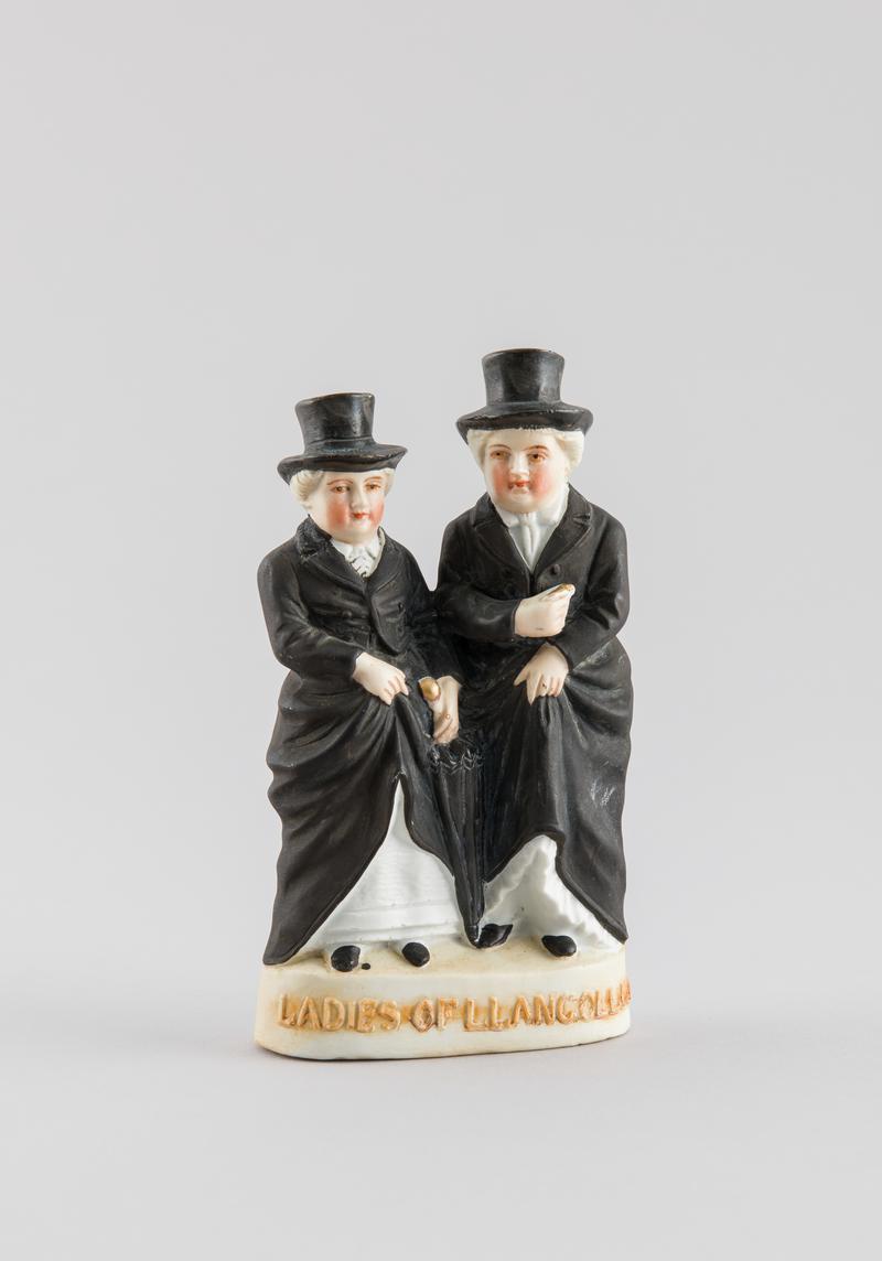 A hollow figurine of two women wearing black with a black top hat, on the front of the base are the words LADIES OF LLANGOLLEN.