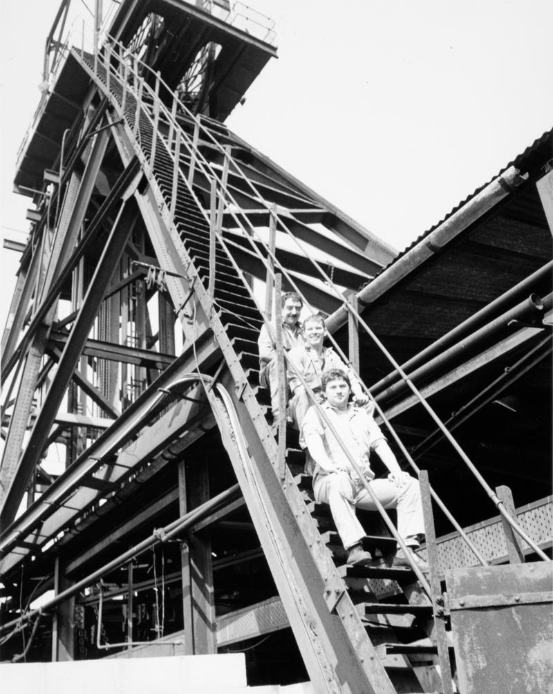 Three workmen sitting on the steps of winding gear of an unknown colliery.