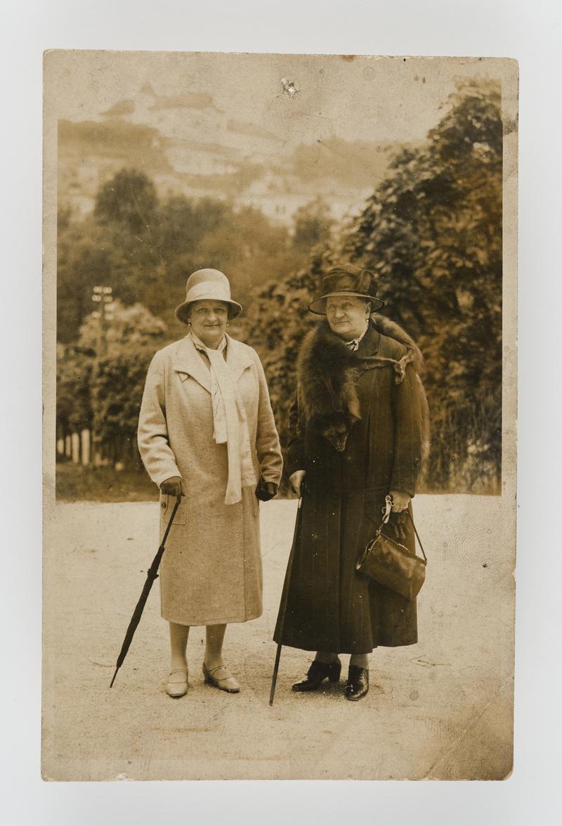 Outdoor photograph of two women in coats and hats (one in furs with walking stick; the other with a rolled umbrella).