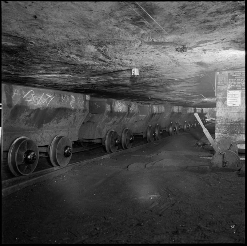 Black and white film negative showing a line of drams in the Graigola Seam, Graig Merthyr Colliery.  Note the unsupported sandstone roof.