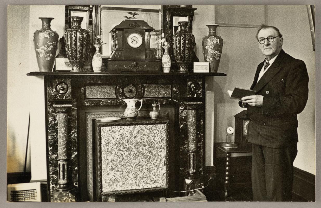 View of C.H. Watkins standing holding a book in front of fireplace at home.