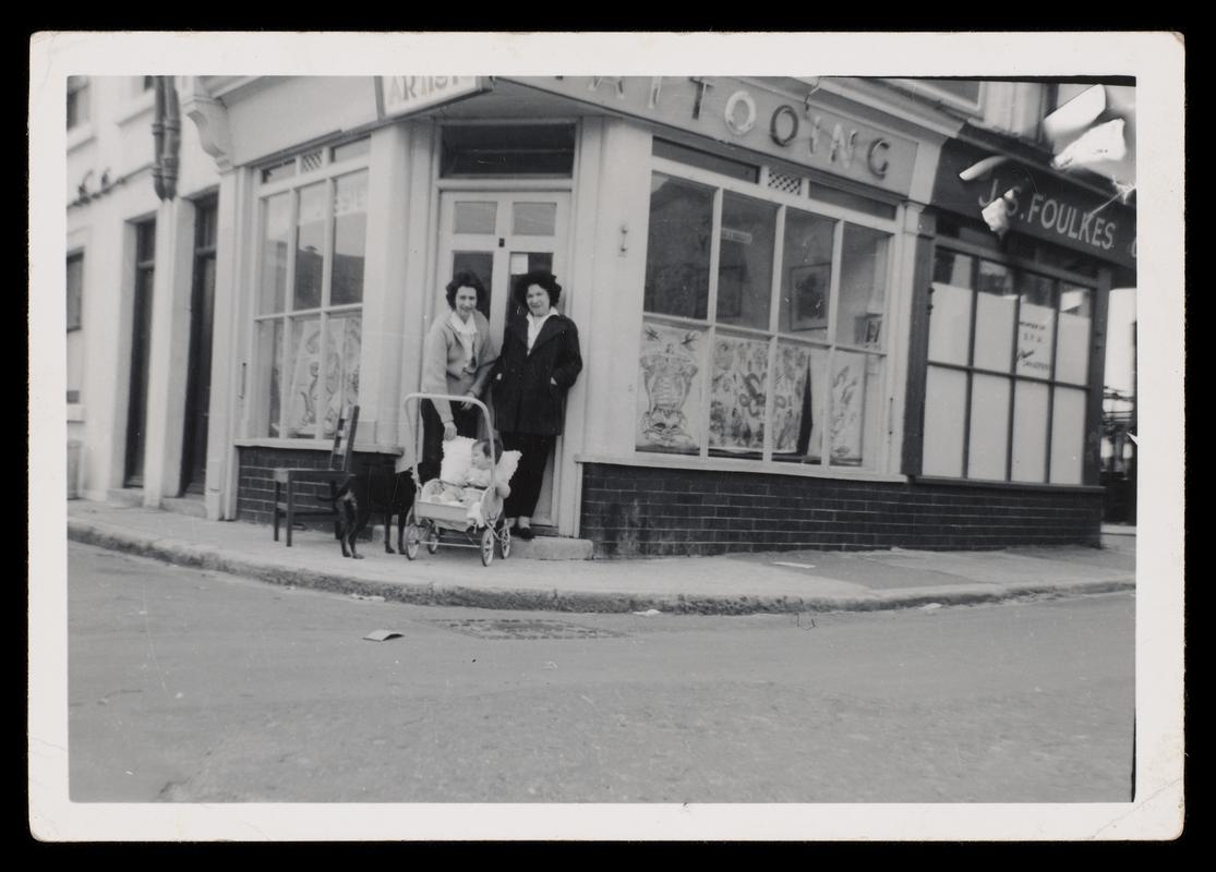 Black and white photograph of two females, baby in pram and dog outside tattoo shop.