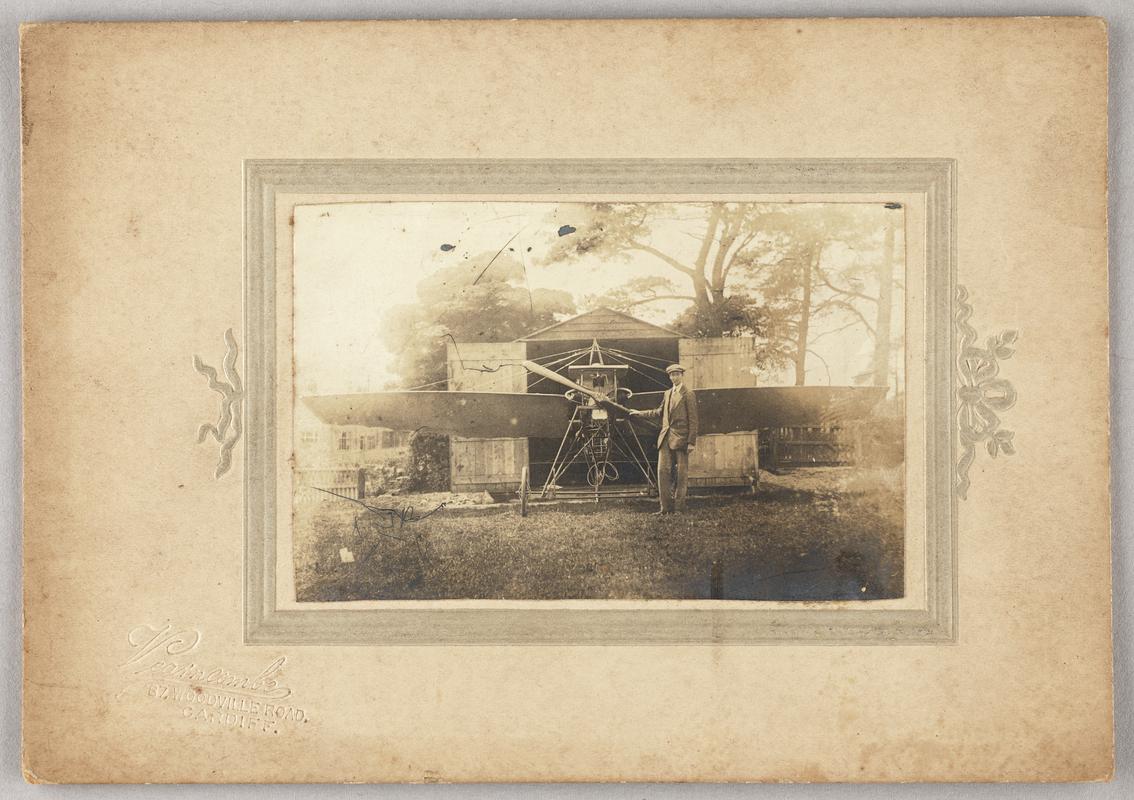 C.H. Watkins as a young man standing next to his aeroplane in front of a shed. Print mounted on card with photographers details printed bottom left of mount.