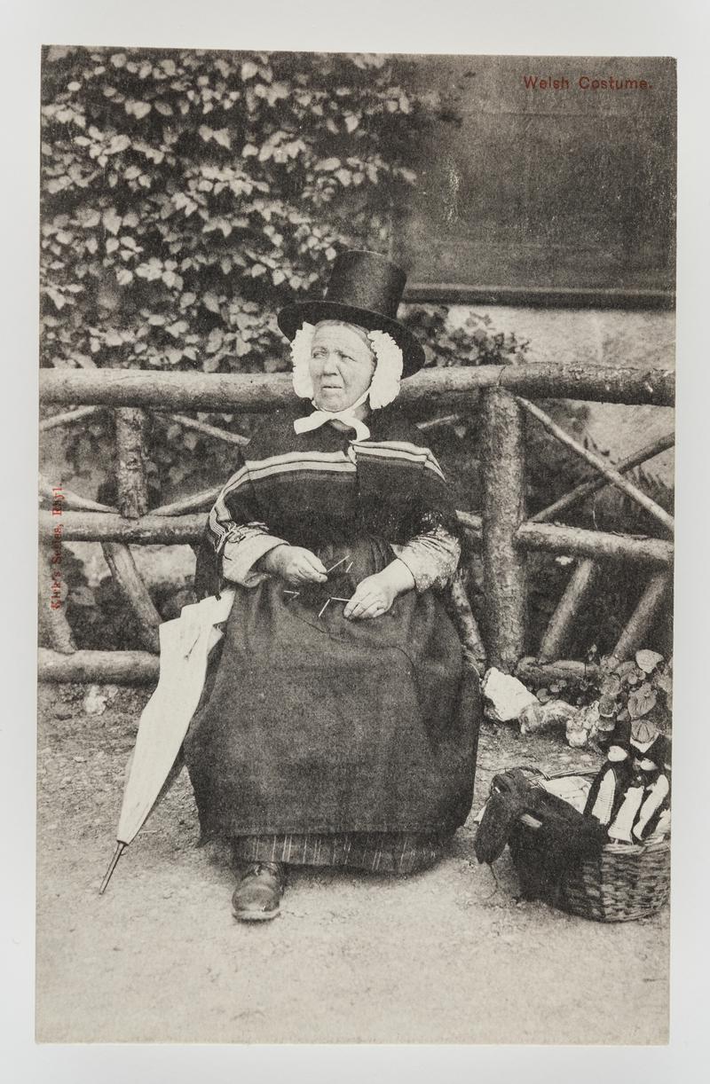 Woman in Welsh costume knitting in garden - umbrella on her arm; wool & Welsh dolls in basket beside her.