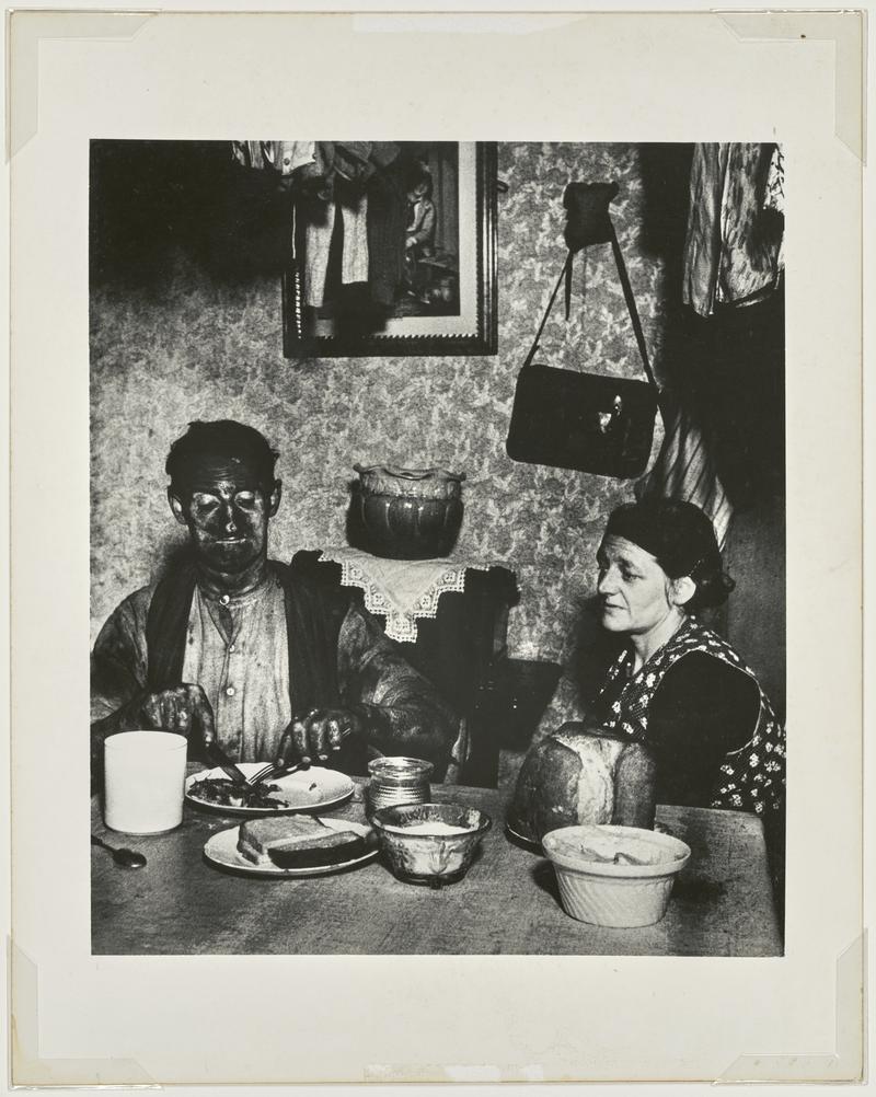 Northumbrian coal miner eating his evening meal, with grubby face and watched by his wife
