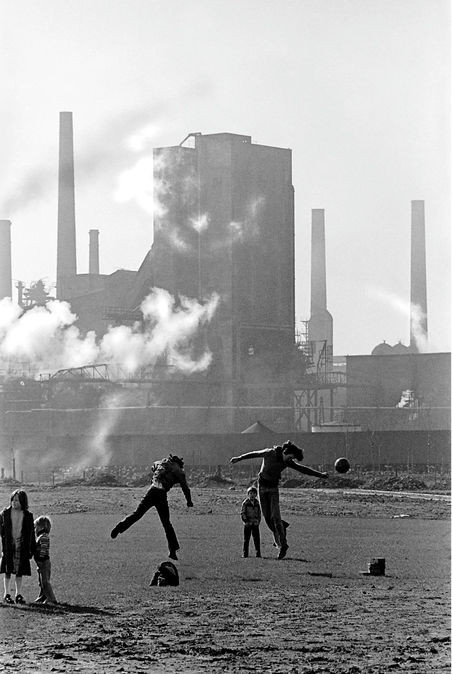 Children play football in spare land in front of East Moors steel works at the time that the steel works were closed. Cardiff, Wales