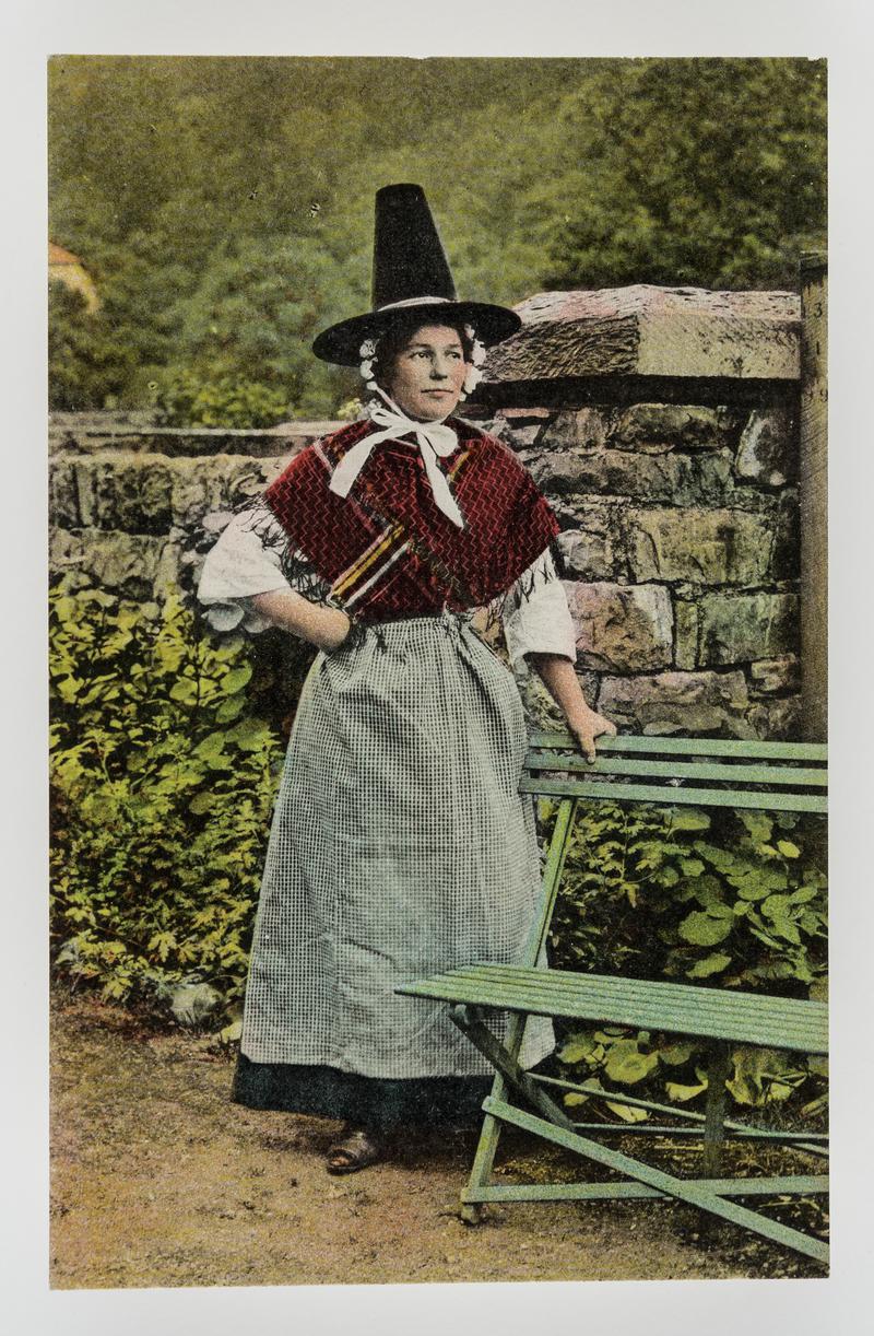 Woman in Welsh costume resting against green bench.