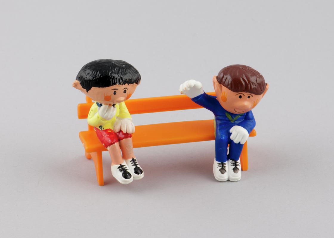 Sitting model of character Basil (2000.179/4) from the Magic Roundabout & Sitting model of character Paul (2000.179.5) from the Magic Roundabout. Model of orange plastic bench (2000.179/14) with two holes on seat for figures from the Magic Roundabout to be inserted.
