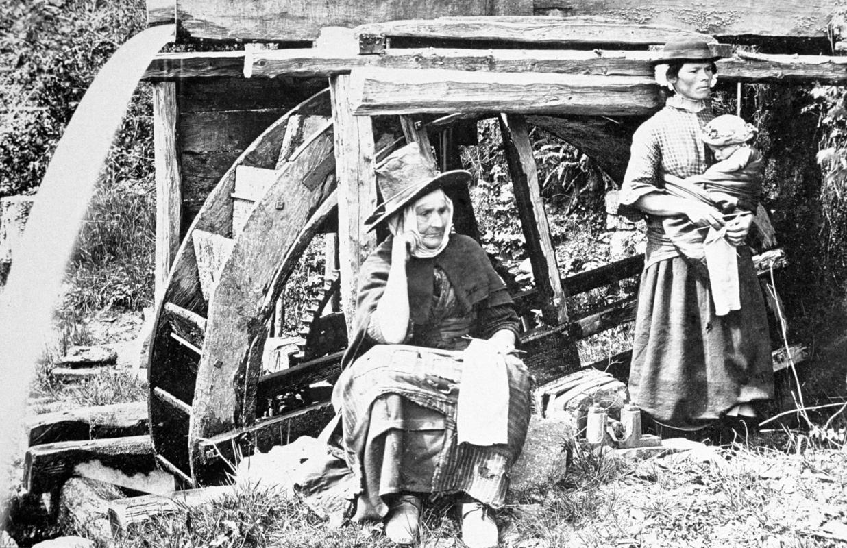 Two women and a baby near water wheel in occupational dress c. 1880 - 1900