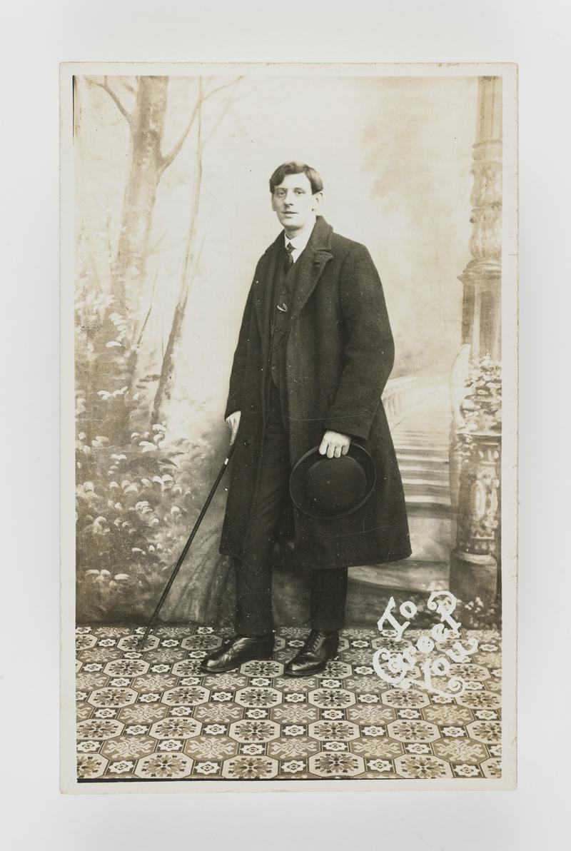 Studio photograph of a young man wearing a suit and overcoat carrying cane and bowler hat.