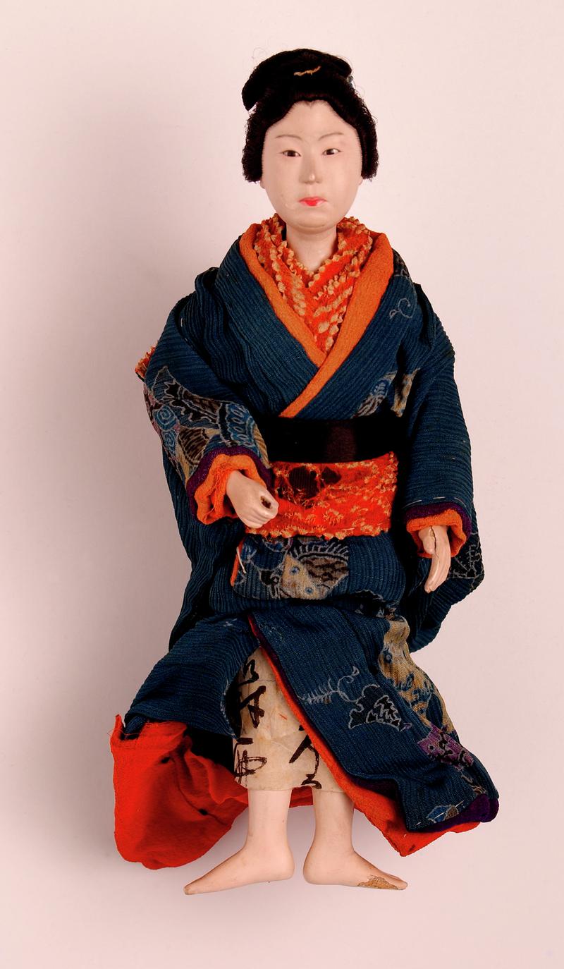 'Sakura' type Japanese doll (standing); head, hands and lower legs made of wood covered with gofum of a fleshy colour; intricate coiffure tied with ribbons; narrow black eyes; right hand is held forward, and probably held a parasol; costume is a patterne