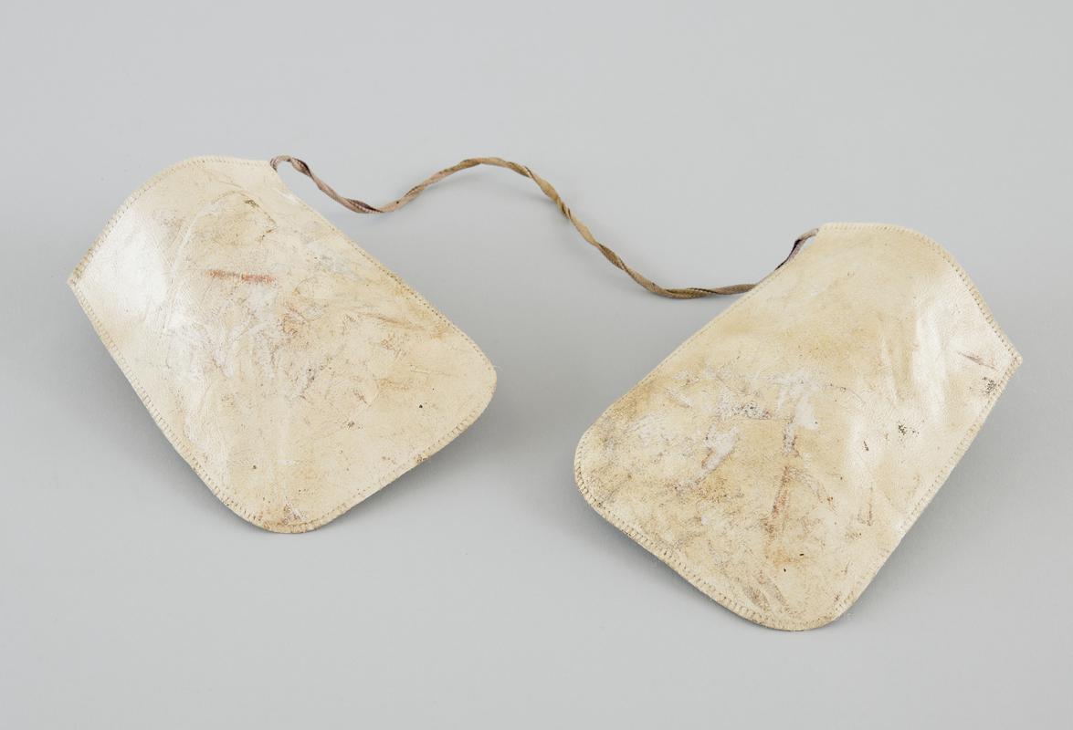Pair of cream oven gloves,  'leatherette' material, attached together with linen strap. Image shows upper side of gloves. Well used, stained and scuffed.