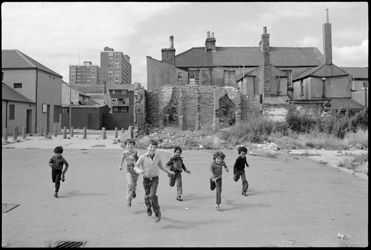 Children in Butetown running towards camera. New and derelict housing, including Loudoun Square flats, in the background.