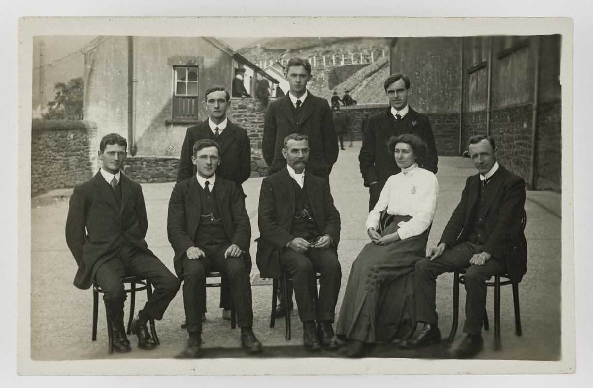 Group of 7 men and 1 woman, in (?) school playground. (Possibly teachers.)