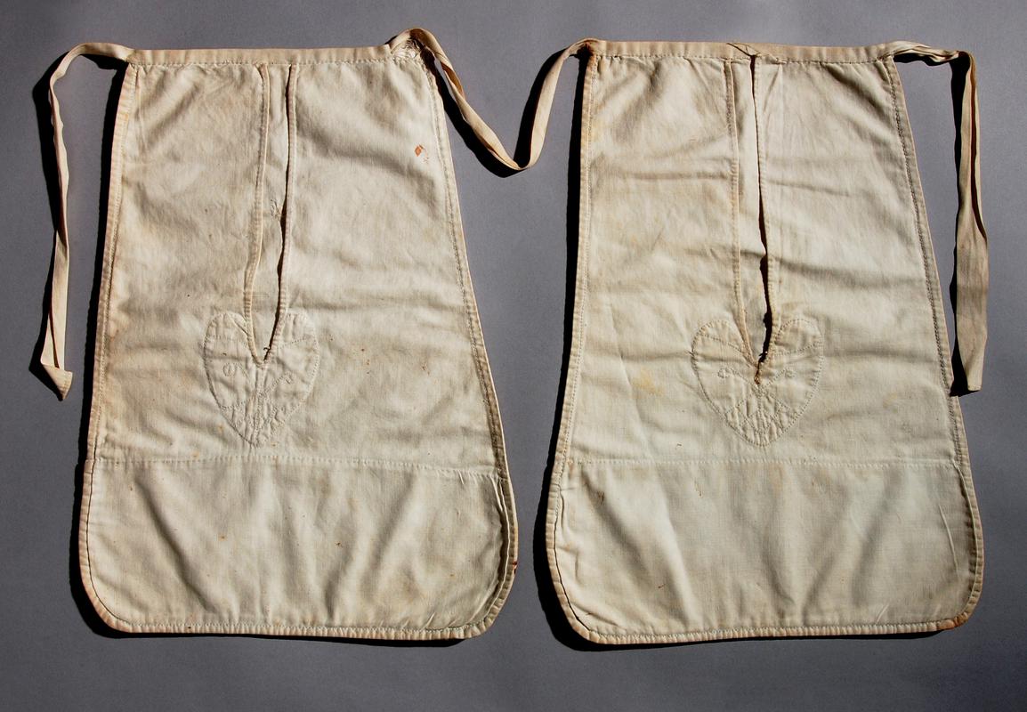 Pair of white cotton twill pockets; tape joining pockets and tape ties at waist.  All hand stitched.  Pocket openings turned over and tacked down with running stitch; base of openings decorated with a heart shaped motif in white cotton backstitch.  (Photographed for Winchester project.)