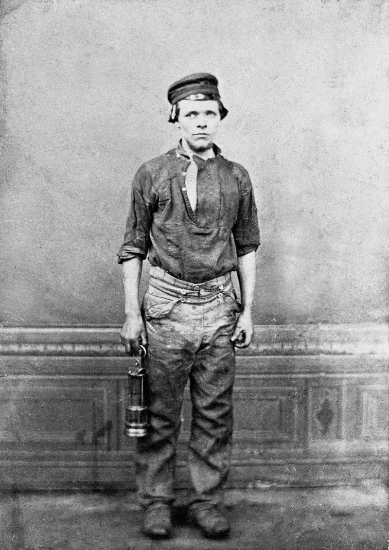 Colliery worker holding a miner's lamp in right hand.