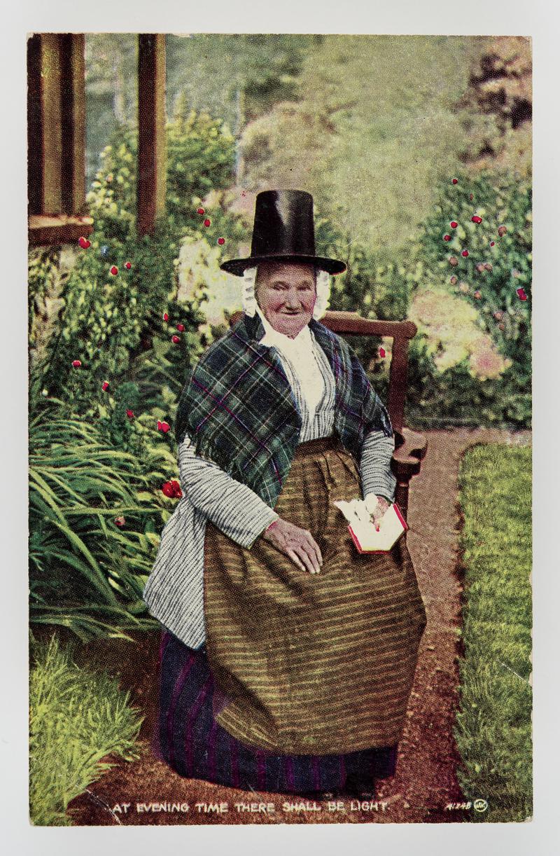 Elderly lady in Welsh costume sitting in garden with book. Inscribed 'At evening time there shall be light.'
