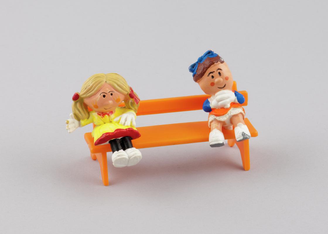 Sitting model of character Rosalie (2000.179/6) from the Magic Roundabout & Sitting model of character Florence (2000.179/7)  from the Magic Roundabout.Model of orange plastic bench (2000.179/14) with two holes on seat for figures from the Magic Roundabout to be inserted.