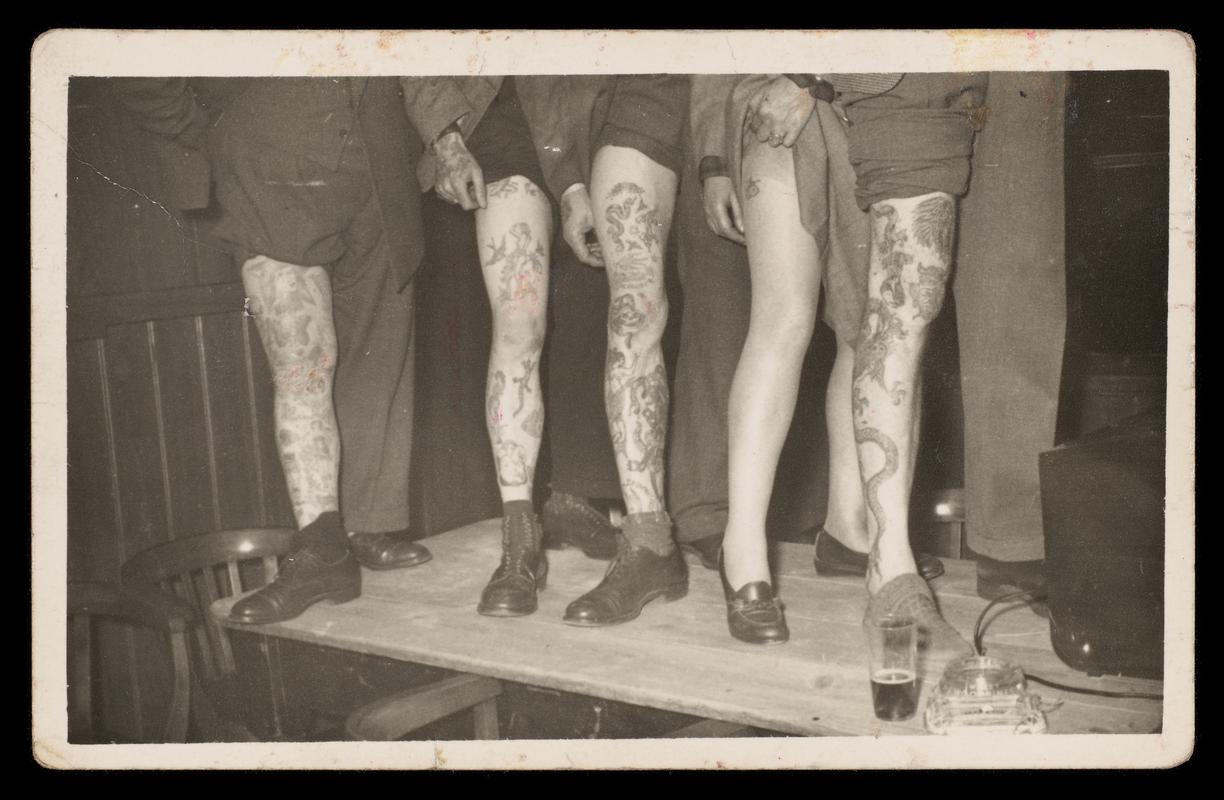 Black and white photograph. Lower half shot of group with tattooed legs