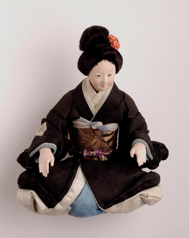 'Sakura' type Japanese doll, in kneeling position; head, forearms and lower legs of wood coated with gofum of a slightly fleshy colour; intricate coiffure, with artificial flower and tassel; rectangular holes for eyes; costume of black kimono; with usual