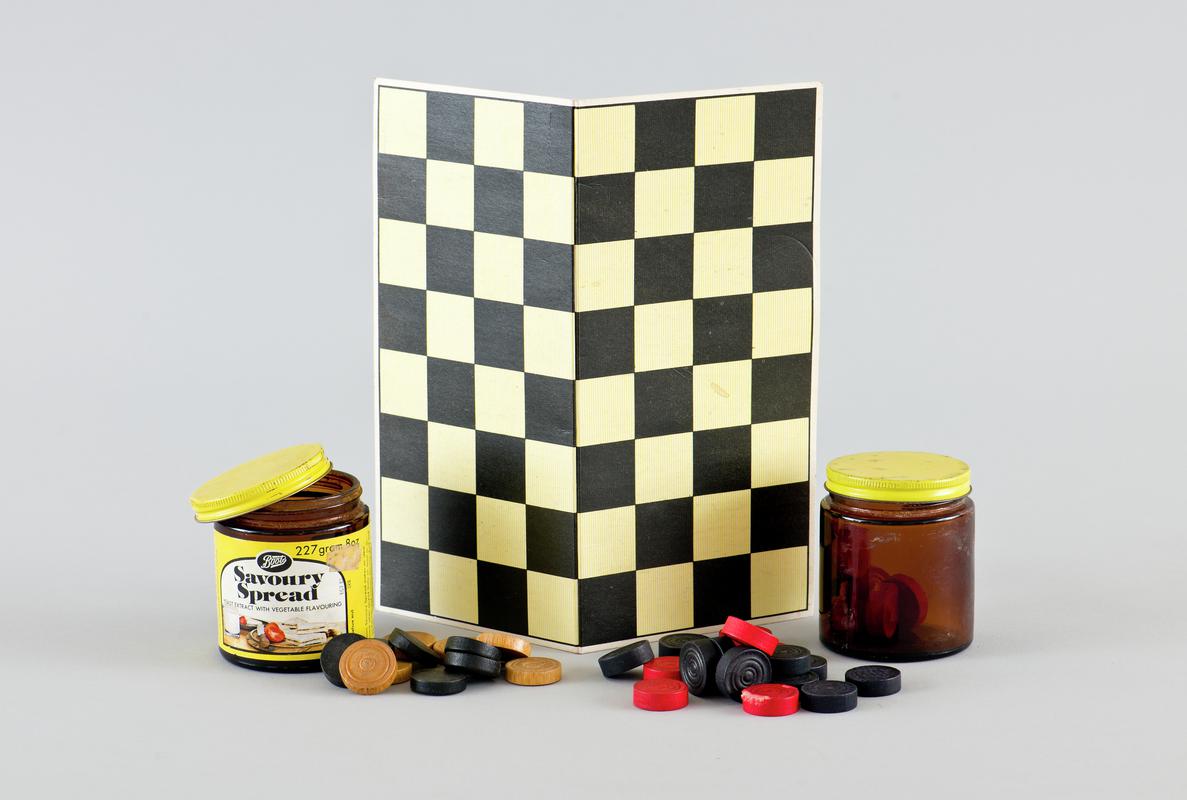 Cardboard draughts board, and two 8oz 'Boots Savoury Spread' jars containing two sets of draught pieces: one set of black and tan draughts (11 of each) and one set of black and red draughts (12 of each and some counters damaged).