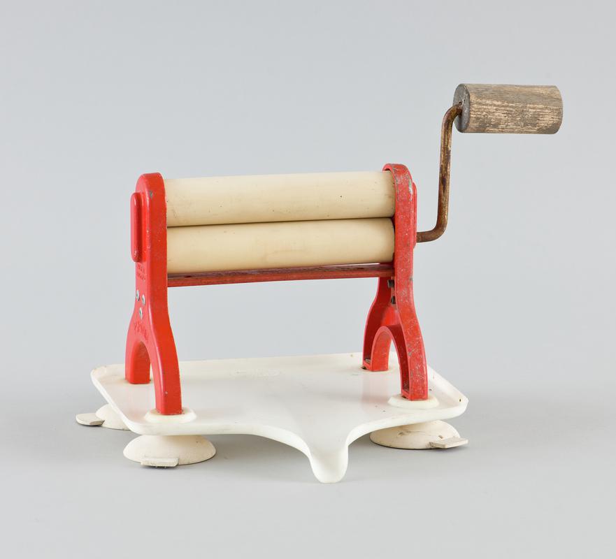 Tabletop clothes mangle called the 'Portair Limpet'. Cream rubber (?) rollers within a red metal frame with well worn wooden handle to operate, sitting in a white plastic water tray with spout that can direct water into bowl. Tray sits on four cream rubber feet that can be stuck to flat surface. Product title embossed on side of two red metal uprights holding rollers.