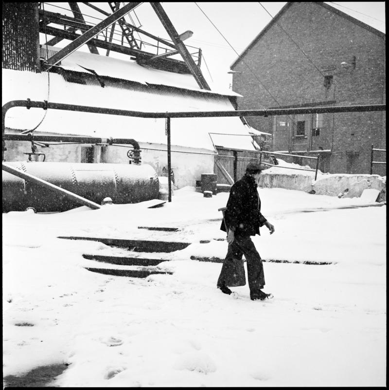 Black and white film negative showing a man walking across the yard, headframe and engine house in the background, Big Pit Colliery.