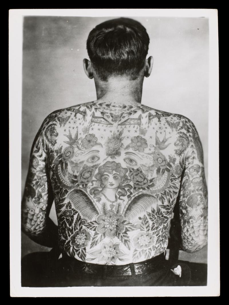 Black and white photograph: half figure of male with back tattooed.