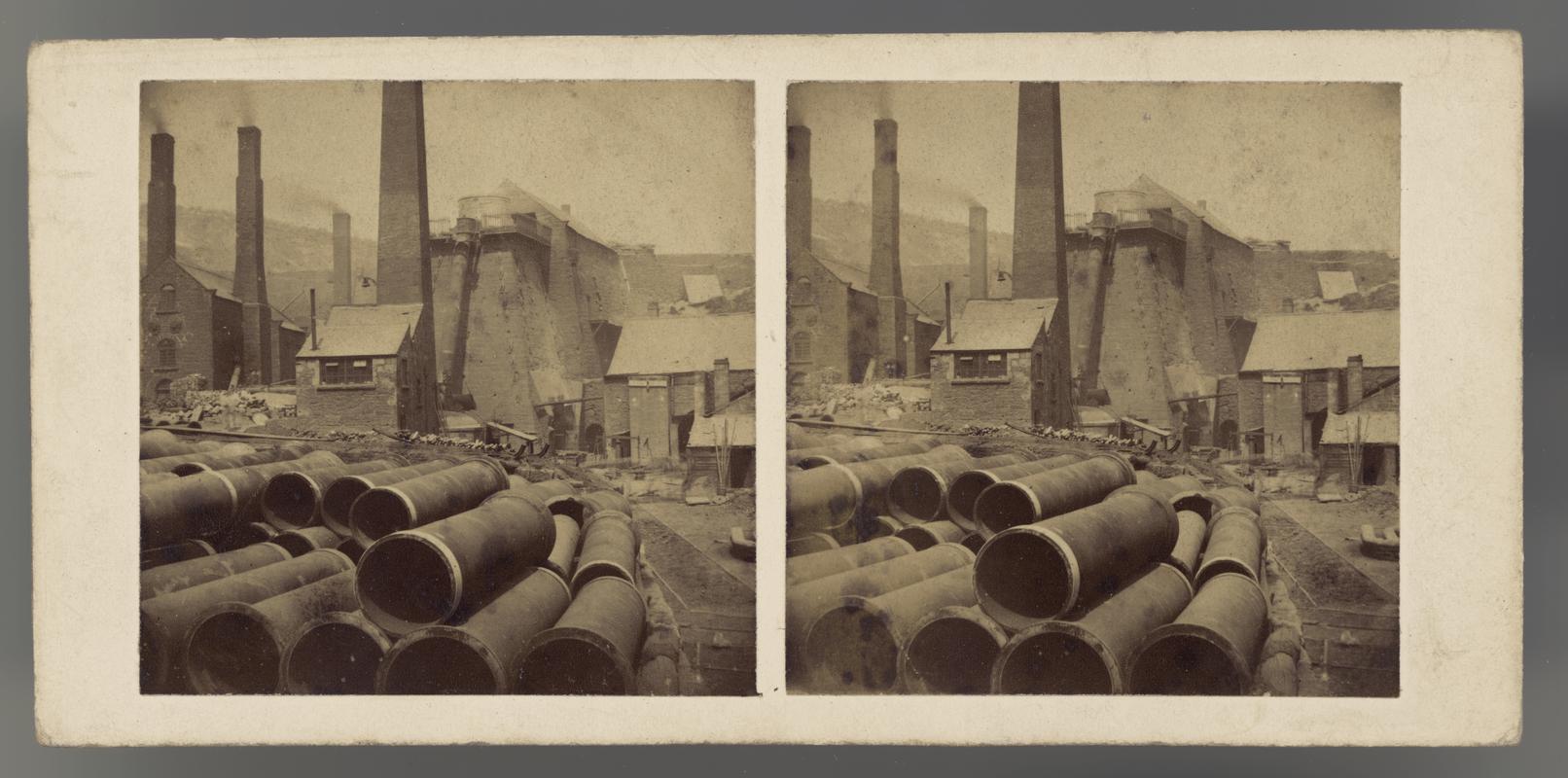 Stereoscopic photo card showing stacks of pipes with a blast furnace in the background, Brymbo.