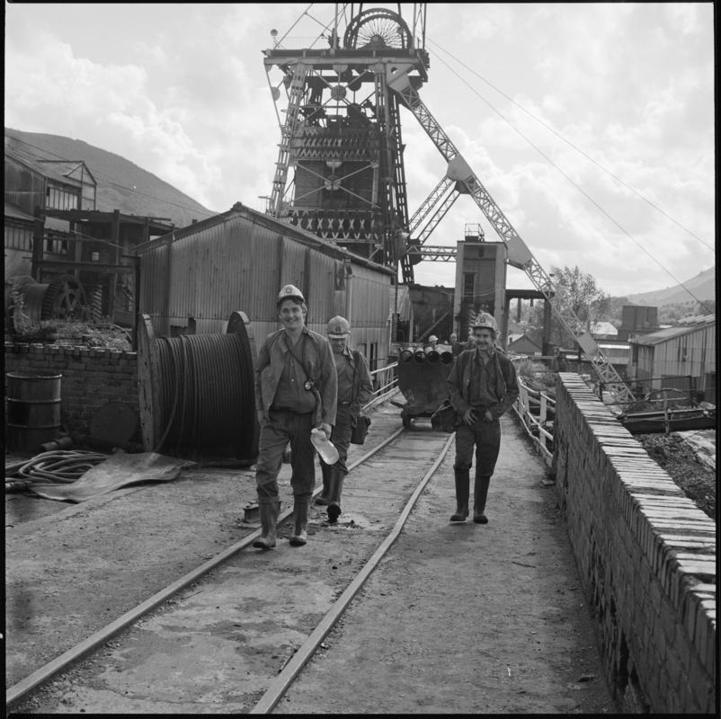 Black and white film negative showing a surface view of Merthyr Vale Colliery with three men in the foreground, 1976.  'Merthyr Vale 1976' is transcribed from original negative bag.