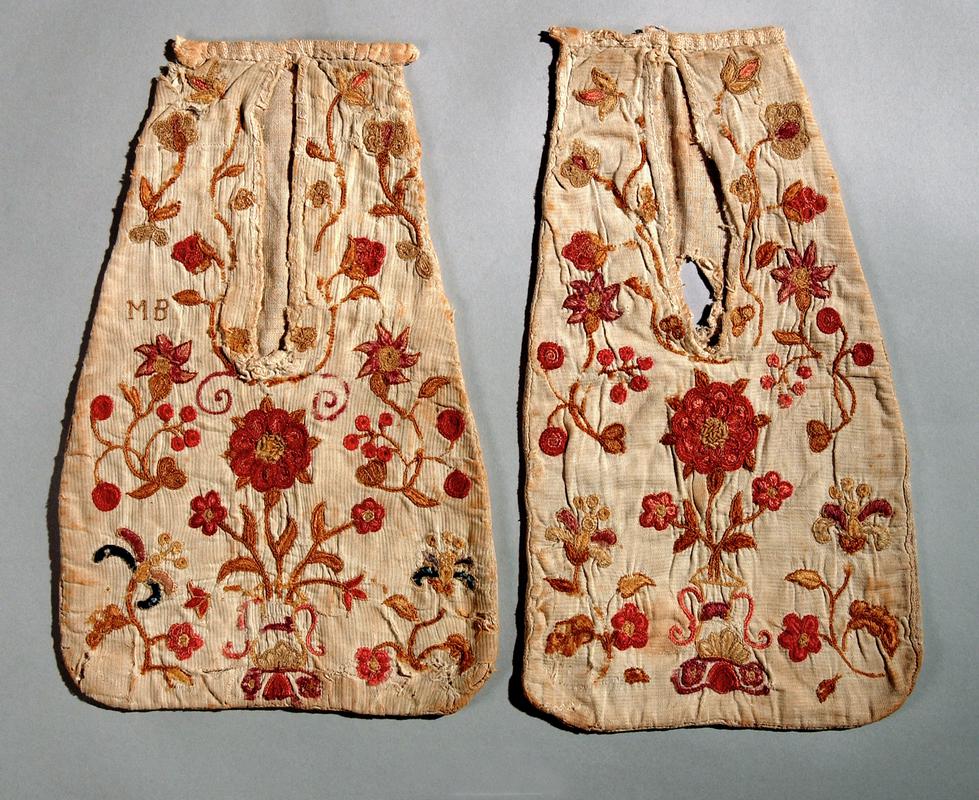 Pair of pockets in fancy weave white cotton; plain weave backing.  Fronts embroidered overall in fine wools with flower, rose and leaf motifs in pink and yellows, (some blue on /15 and initials MB) in chain and stem stitch. Tops bound with coarsely woven tape; fastening ties cut off.  Late 19th century. From Capel Bangor, Ceredigion.  (Photographed for Winchester project.)