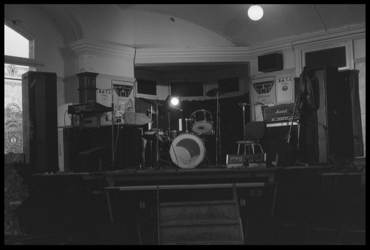 TNT Group's musical instruments on stage in corner of room. Location unknown.