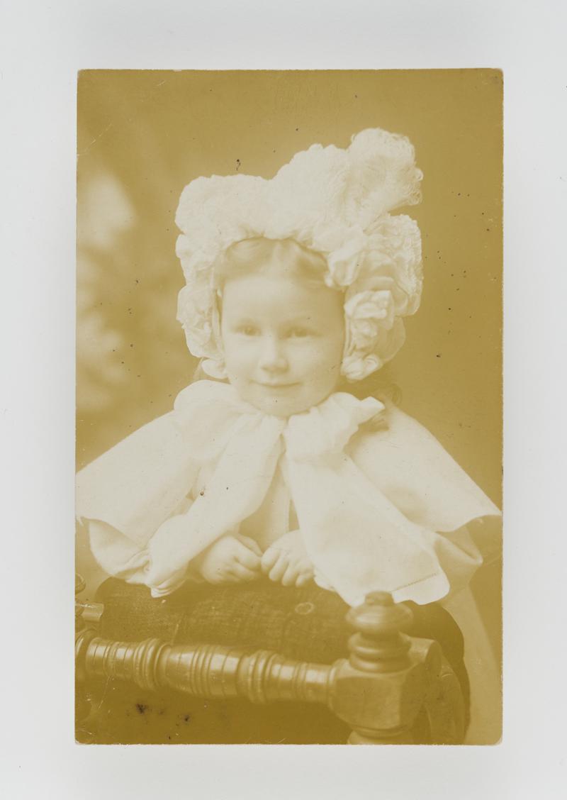 Head & shoulders photograph of a young girl in bonnet looking over chair. (1900s)