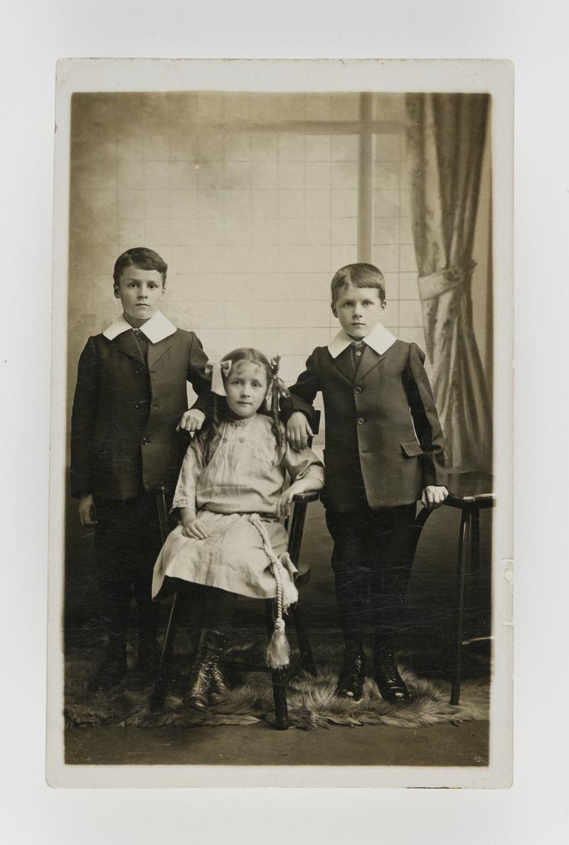Indoor photograph of two young boys standing wearing matching suits with a young girl sat in between them.
