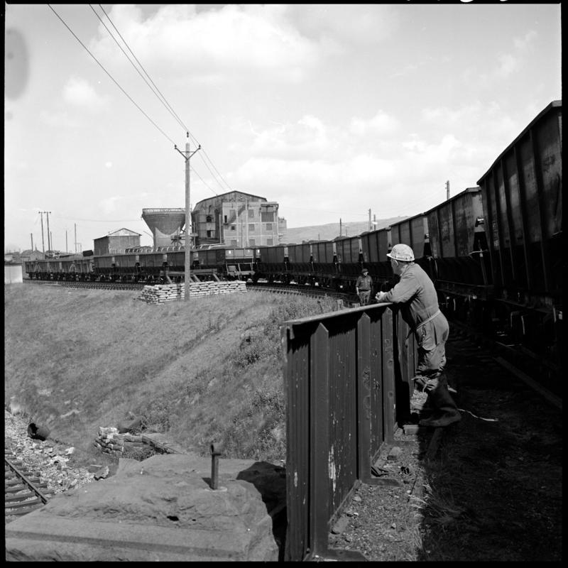 Black and white film negative showing a locomotive passing the Big Pit washery, with a man leaning on fence in the foreground.