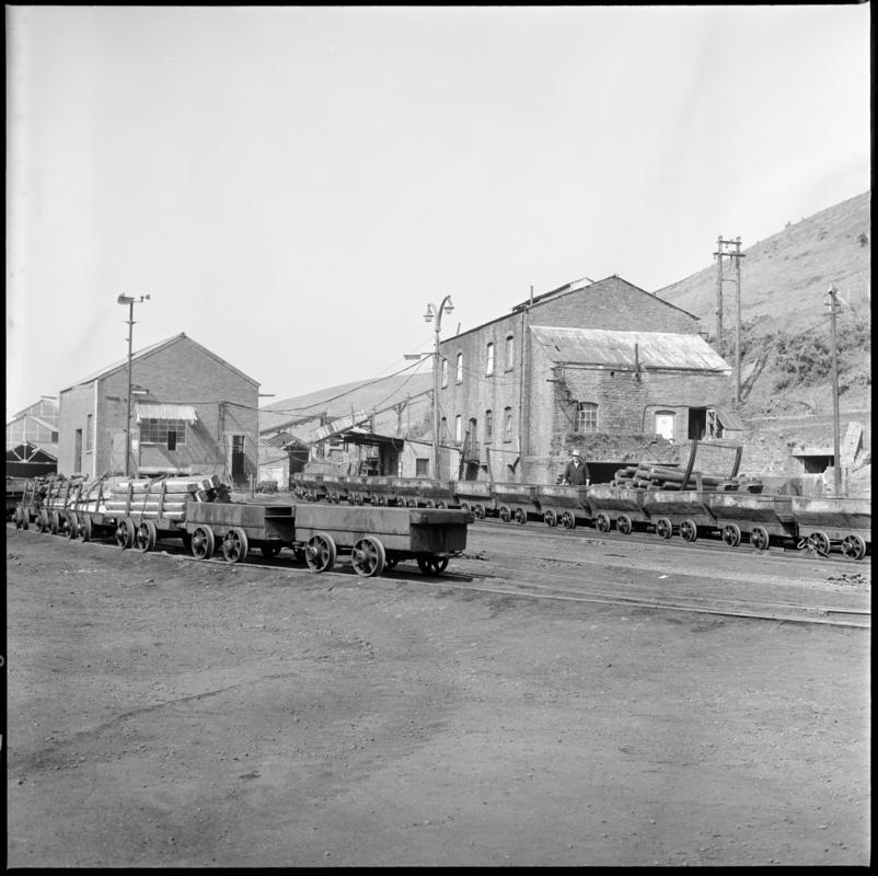 Black and white film negative showing Graig Merthyr Colliery yard in 1977 with a long journey of drams waiting to be run into the mine.  In the foreground are special cut-down drams used for moving supplies.  'Graig Merthyr' is transcribed from original negative bag.