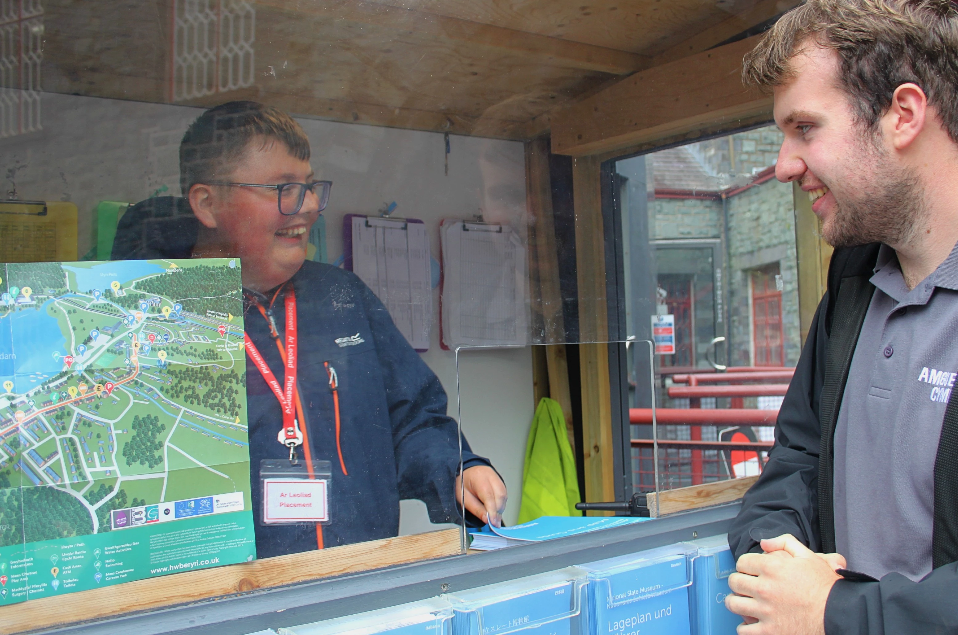 Unigolyn yn sefyll yn y cwt croeso yn yr Amgueddfa Lechi, yn gwenu wrth siarad â aelod o staff. 
