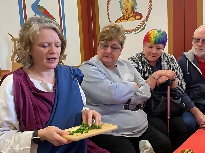 Menyw wedi gwisgo yn dillad Rhufeinig yn trafod defnyddiau llysieuol gydag ymwelwyr