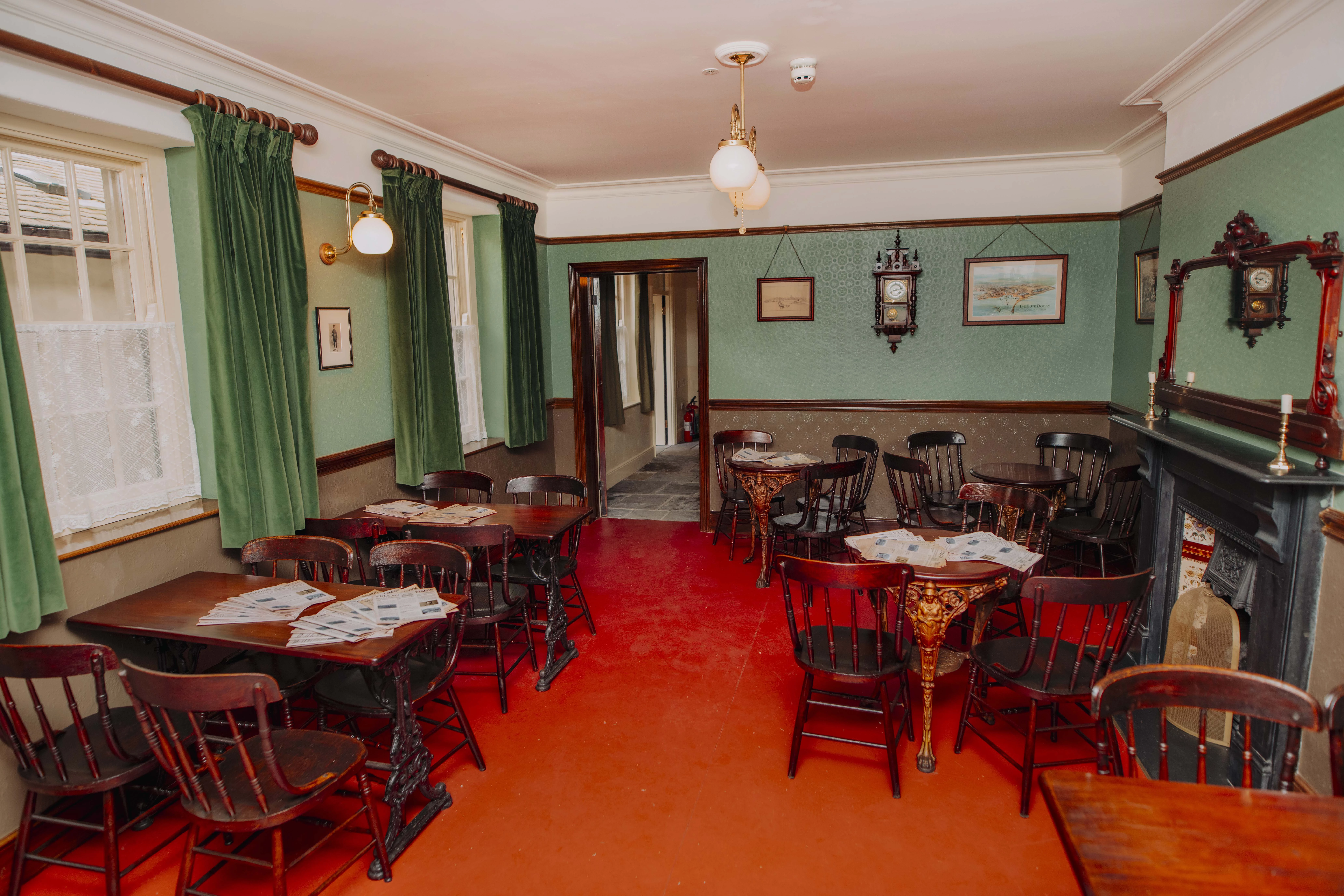 Ystafell glyd gyda llawr coch, waliau a llenni gwyrdd, byrddau a chadeiriau pren, lle tân a lluniau wedi'u fframio ar y waliau.