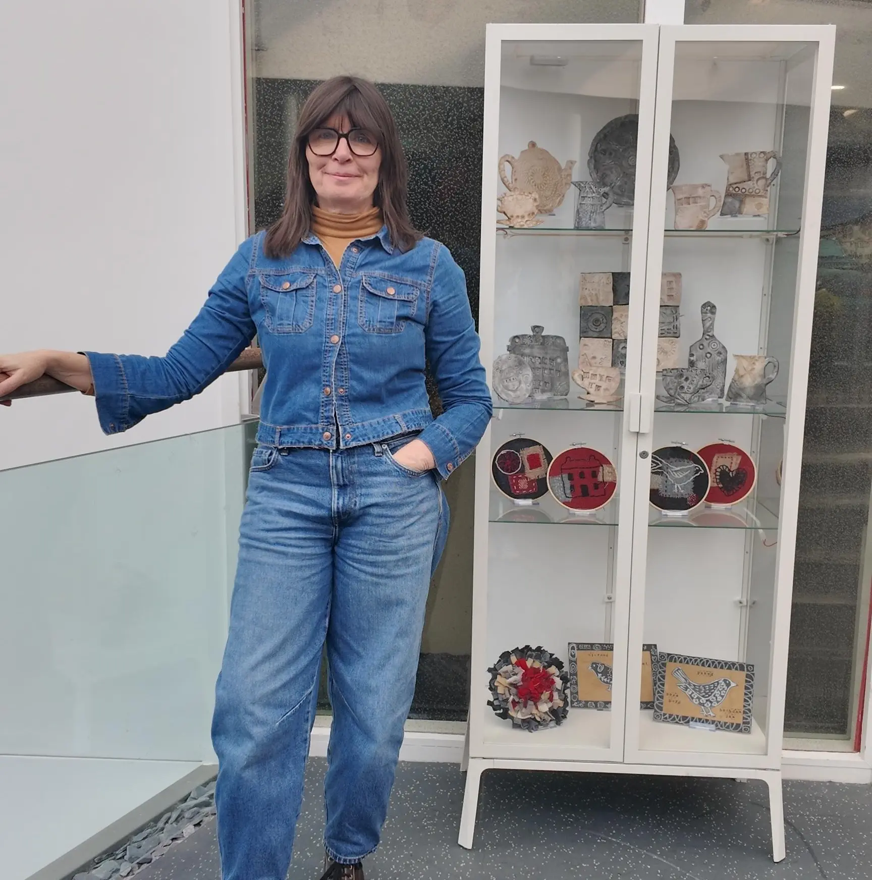 Artist, Rachel Evans, yn sefyll wrth arddangosiad o weithiau creadigol wedi greu during gweithdai yn Ysbyty Alltwen. 