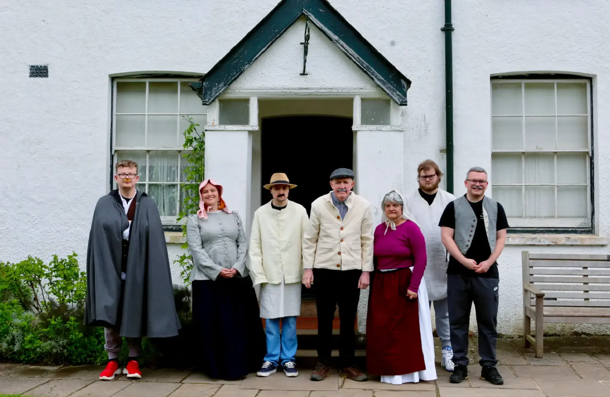 Pobl mewn cymysgedd o ddillad hanesyddol a modern yn sefyll gyda’i gilydd y tu allan i dŷ fferm gwyn traddodiadol.