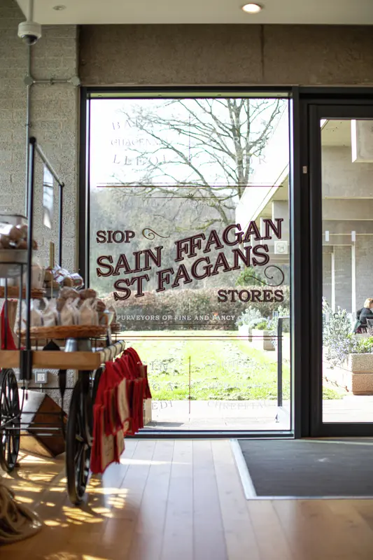Ffenestr siop gyda llythrennau 'St Fagans Stores', yn edrych allan ar ardd heulog gyda char pren o flaen.