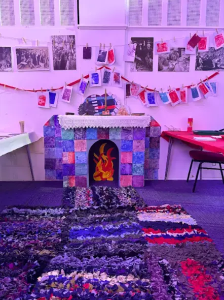Lle tân wedi'i greu o gardboard yn liwgar gyda gwrthrychau o'r amgueddfa ar y silff uwchben, matiau rhacs a phrintiadau ynghyd â lluniau o deuluoedd wedi'u gosod ar y wal yn creu profiad ac awyrgylch ystafell fyw.